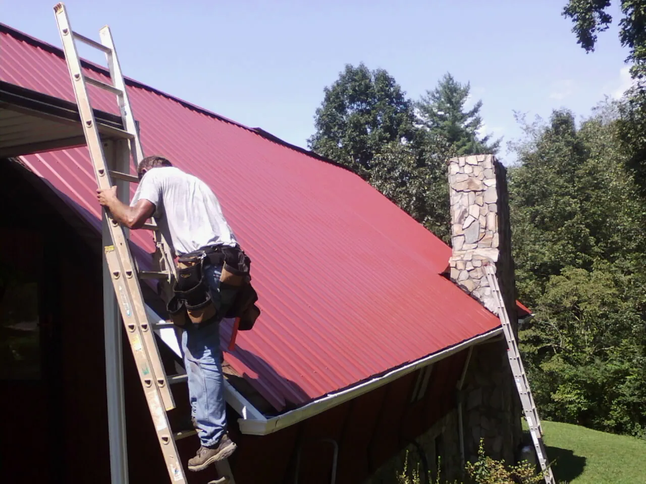 Roofer on a ladder beside a red metal roof and stone chimney — Soffit & Fascia Repair by Roofing Brothers in Mount Vision